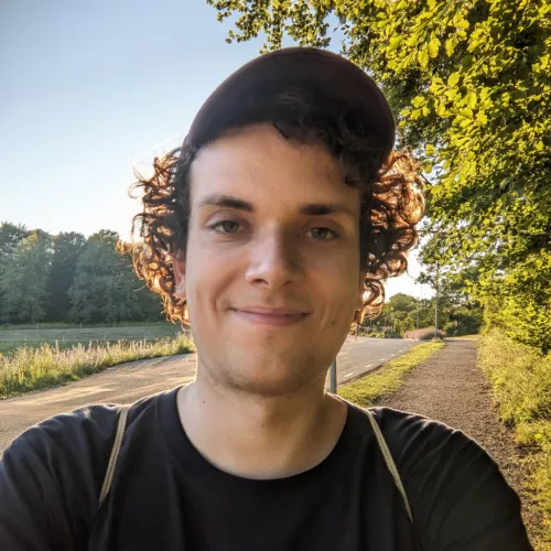 Eine Person mit Locken und Cappy auf dem Kopf und dunklem T-Shirt schaut in die Kamera. Im Hintergrund sind Wiesen und Bäume zu sehen und goldener Sonnenschein.
