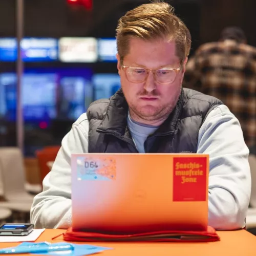 Ulrich Berger arbeitet konzentriert an einem Laptop, auf dem ein D64-Sticker und ein roter Aufkleber "Faschismusfreie Zone"; zu sehen sind. Launch des Code of Conduct Demokratische KI, 2025.